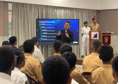 Presenter in a black shirt explains a slide titled 'Consequences of Road Accidents' to uniformed students in a hall.