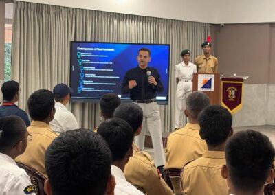 Speaker in a dark shirt with a microphone presents to cadets while a slide on road accidents is shown on a large screen behind him in a hall?