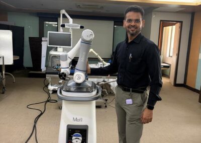 Man in black shirt and khaki pants standing beside a Meril robotic surgical system in a clinic, with cables and monitor nearby