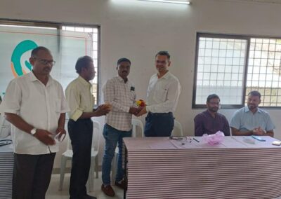 Office ceremony: two men exchange a small bouquet as others watch, with a striped table and a banner/logo in the background.